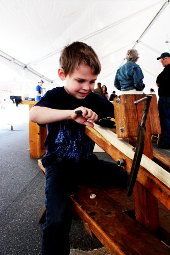 Atticus Cunningham, 6, sits atop a shaving horse to work on a wagon spoke. The tool in his hand is called a “spoke shave.”