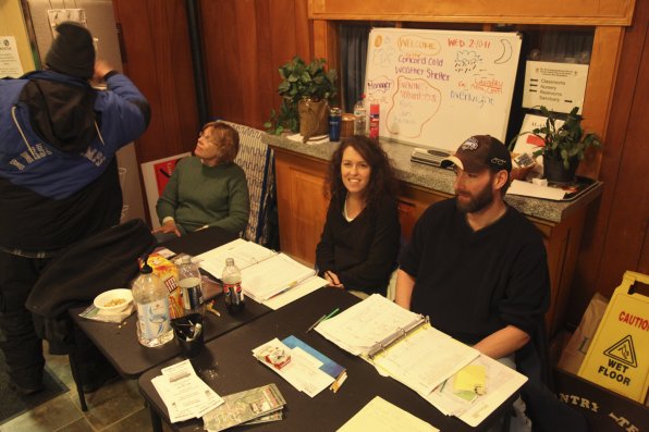 From left, volunteers Barbara Lanweher, Jennifer Kimball,  and Mark Edgecomb, all of Concord, sit at the door and greet residents of the shelter last week. There are 150 volunteers who sign up to work two shifts each night; one is from 5:30 to 9:30 p.m.; the other is 9 p.m. until 8 a.m., when the shelter closes for the day.
