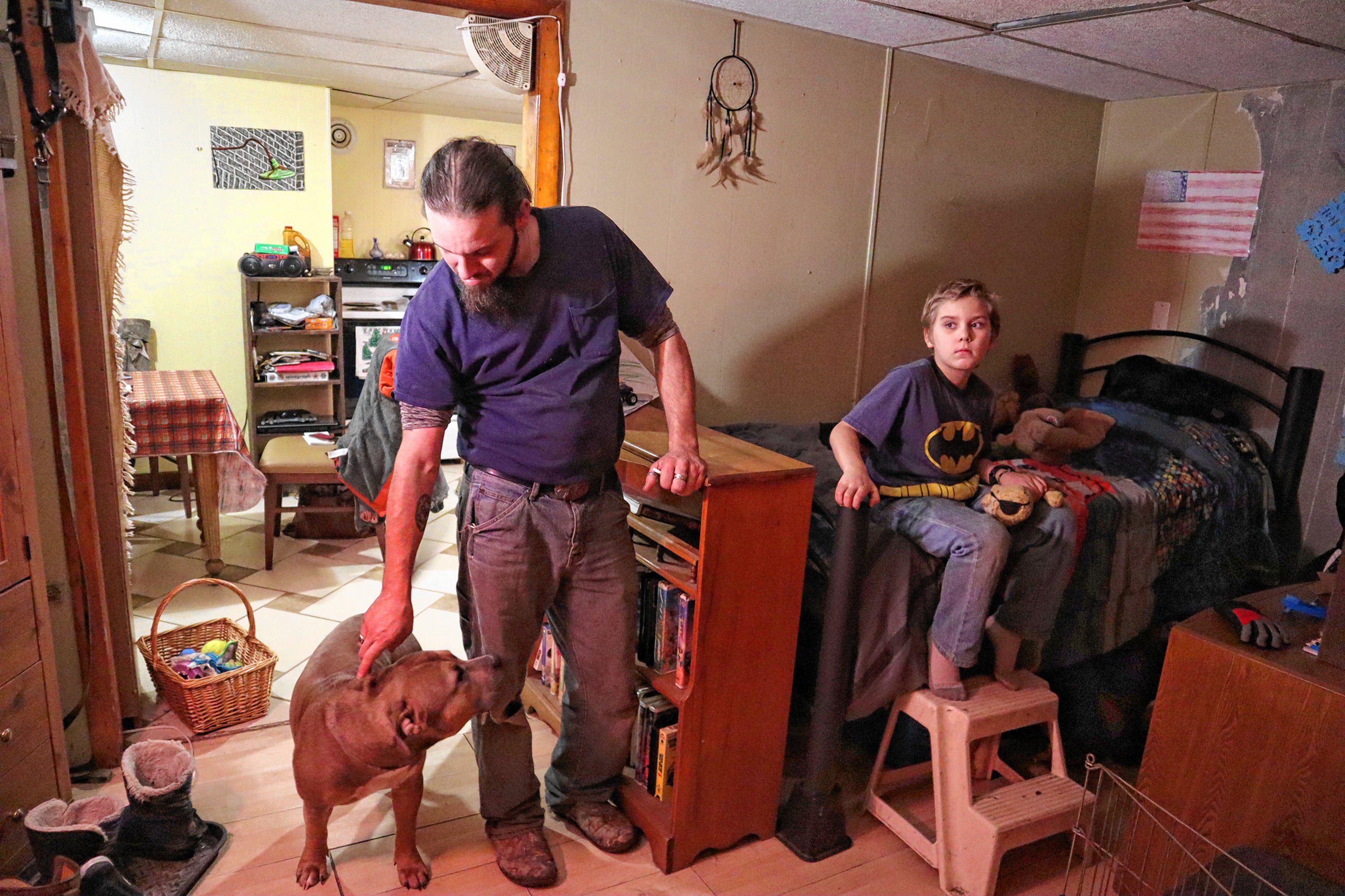 Mark Aucoin leans down to pet his dog, Max, while his 9-year-old son Shaye sits on his bed in the family’s new Chichester apartment. 